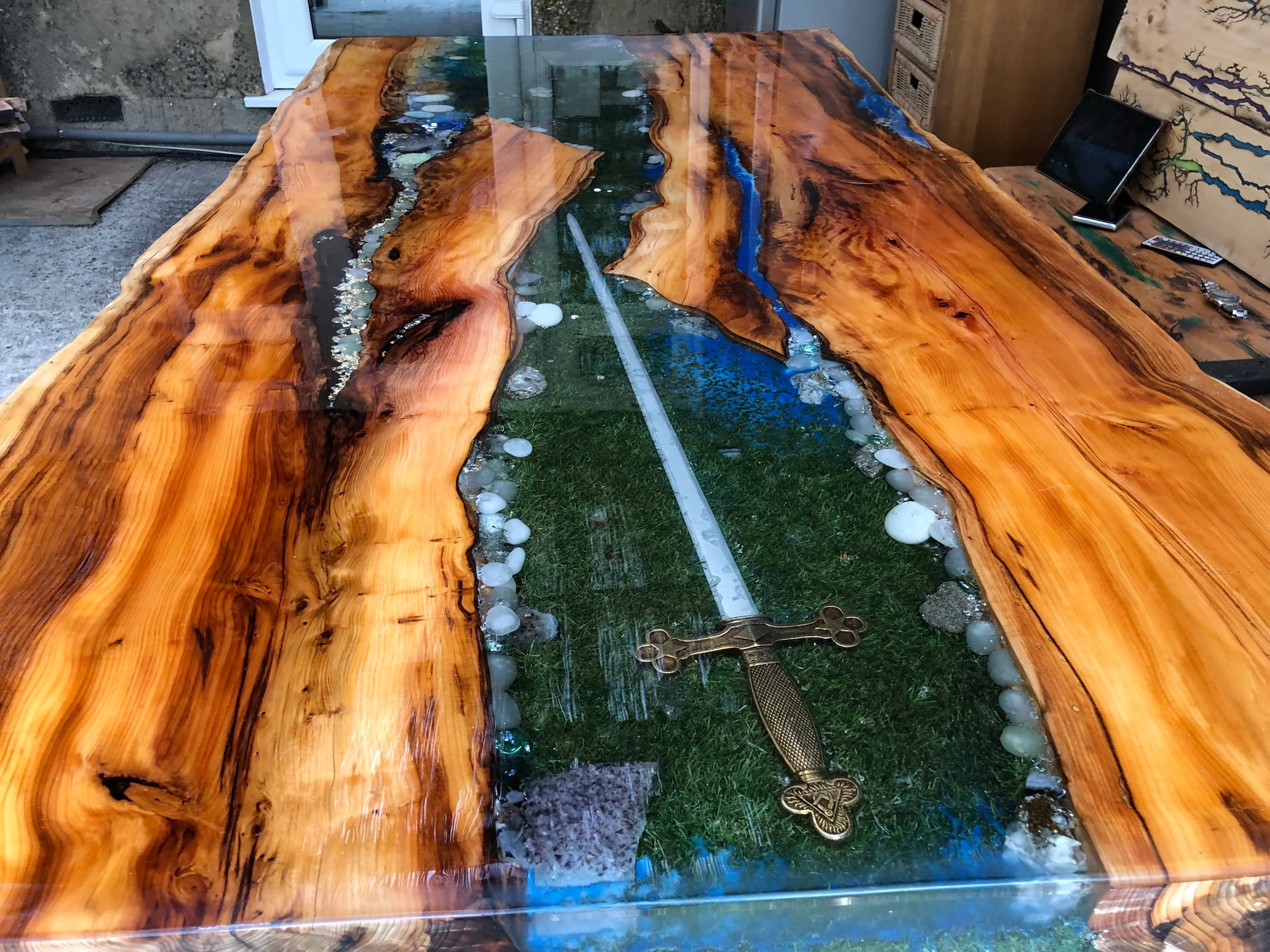 Full ornate sword embedded across a moss-and-resin river through a live-edge tabletop, daylight