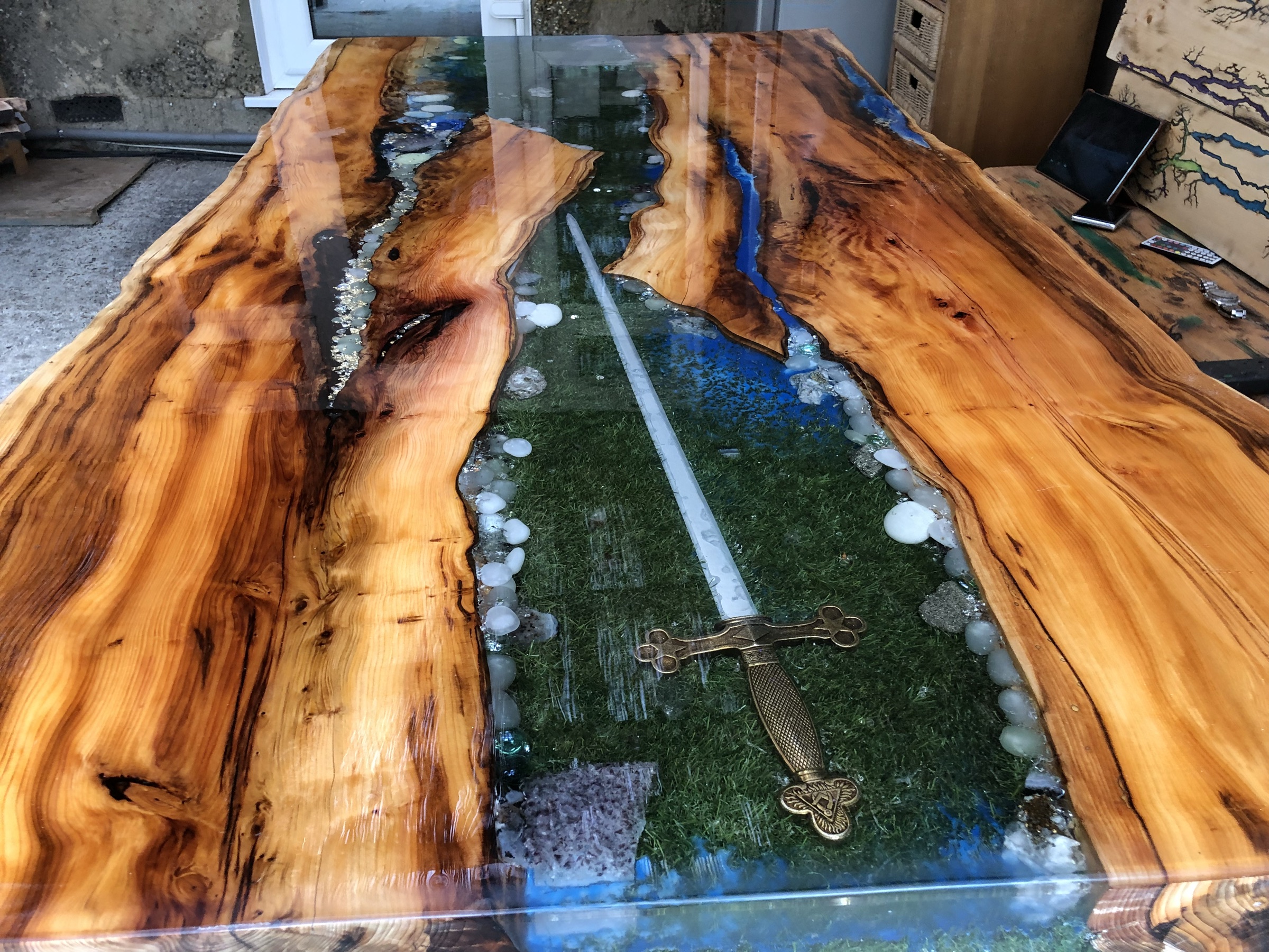 Full ornate sword embedded across a moss-and-resin river through a live-edge tabletop, daylight